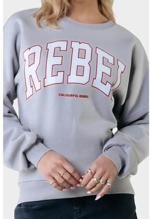 Person trägt einen hellgrauen Pullover mit dem großen roten Schriftzug "REBEL" und darunter steht "COLOURFUL REBEL". Die Hände sind verschränkt.