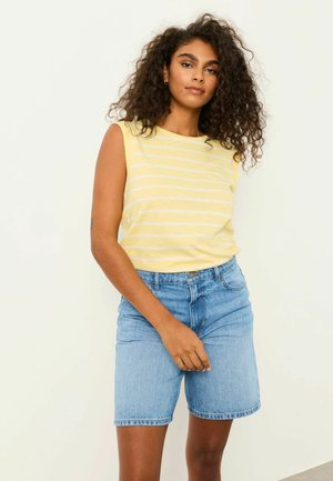 Woman with curly hair wearing a yellow and white striped sleeveless top and blue denim shorts, standing with one hand near waistband.