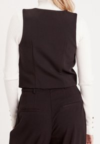 Zwarte mouwloze bodywarmer met een getailleerd ontwerp, gezien vanaf de achterkant. Gemaakt van gladde stof, met subtiele zijzakken en een cropped model.