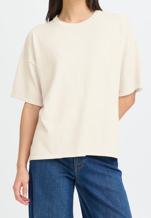 Personne portant un t-shirt ample à manches courtes, uni, blanc cassé, et un jean en denim bleu, debout avec une main détendue sur le côté.
