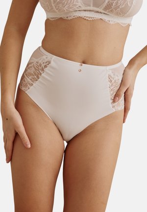 Culotte beige taille haute avec panneaux en dentelle sur les hanches, portée par une personne portant un soutien-gorge en dentelle beige assorti, les mains posées sur les hanches.