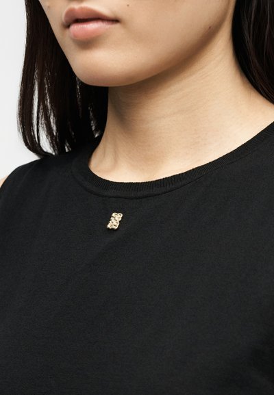 Gros plan sur une personne portant un haut noir avec une petite épingle en forme d'ours en peluche doré près de l'encolure, montrant le bas de son visage et son cou.