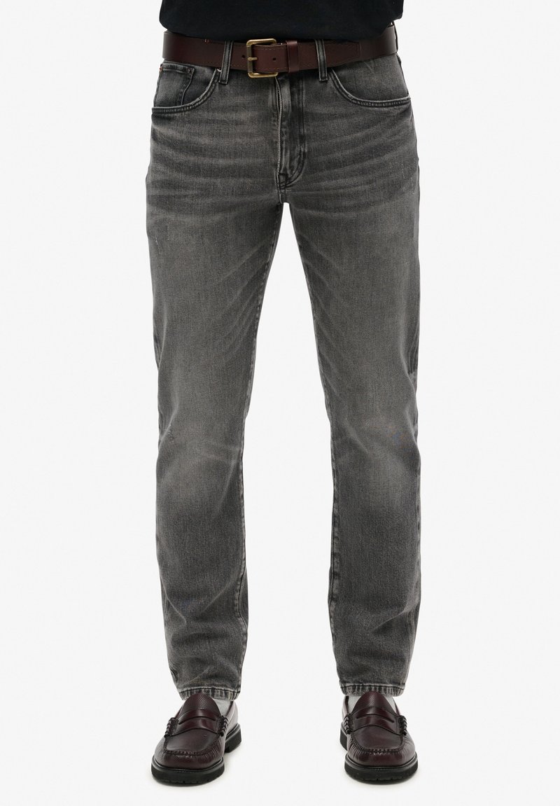 Jeans de denim gris con corte recto, detalles sutiles de desgaste, diseño de cinco bolsillos y un cinturón de cuero marrón. Combinados con mocasines oscuros.