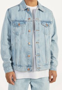 Ljusblå denimjacka med en klassisk design, kopparknappdetaljer, två bröstfickor och ett något texturerat tyg. Lager på en vit skjorta.