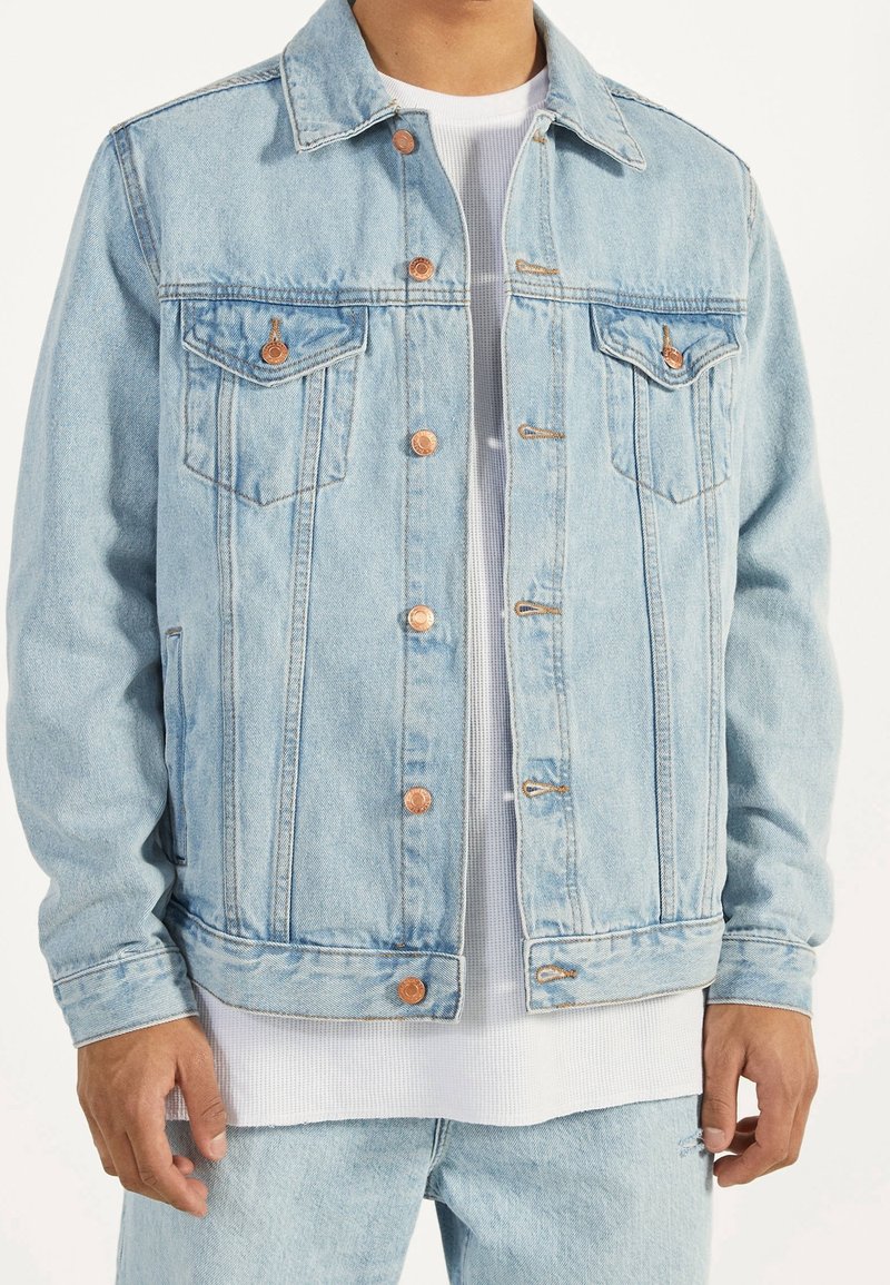 Ljusblå denimjacka med en klassisk design, kopparknappdetaljer, två bröstfickor och ett något texturerat tyg. Lager på en vit skjorta.