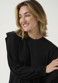 Top negru cu maneci lungi, dintr-un material texturat, cu decolteu rotund striat și maneci încrețite. Prim-plan cu o mână și față, expresie zâmbitoare vizibilă.