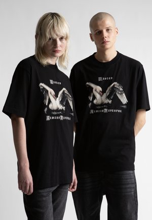 Dwójka młodych dorosłych ubranych w czarne, oversize'owe koszulki z dużą grafiką łabędzia i gotyckim napisem, stojących na tle jednolitego jasnego tła.