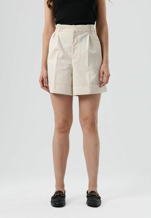 Shorts beige taille haute avec un ourlet replié, des plis devant et des poches latérales. Portés avec des mocassins noirs ornés d'un accent doré.