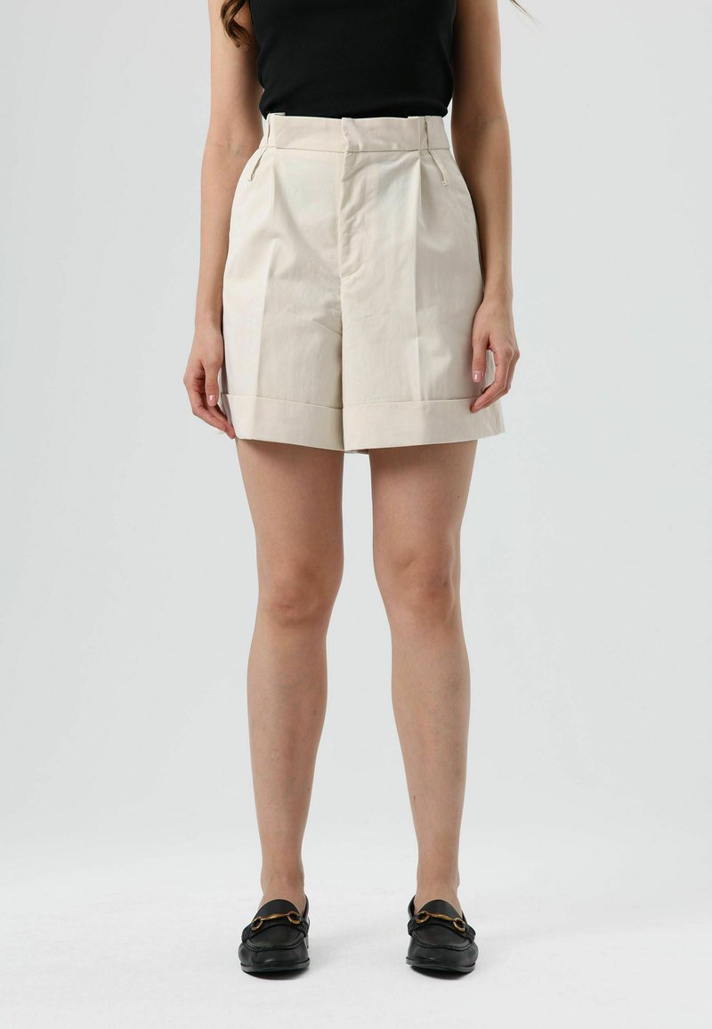 Beige shorts met hoge taille, een omgeslagen zoom, voorplooien en zijzakken. Draagt zwarte loafers met een gouden accent.