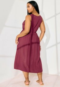 Vestido sin mangas en color burdeos con un cuerpo ajustado y una falda suelta, hecho de una tela ligera, que presenta una cintura fruncida y una textura suave.