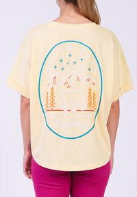 Gele oversized t-shirt met een grafische print van bergen, een oranje beer en de tekst "VERDER ONTDEKKEN" in het blauw.