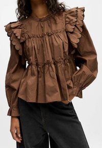 Blouse en coton brun texturé avec un col et des poignets à volants et en scallops ; détail de taille froncée ; manches longues ; associée à un jean foncé.
