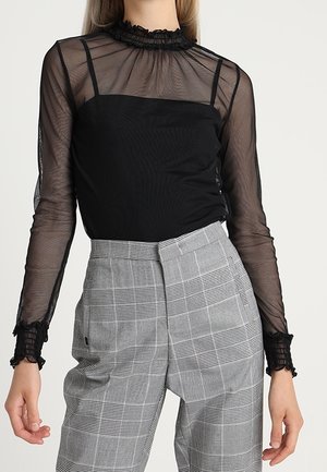 Schwarze transparente Bluse mit einem volanten Ausschnitt und langen Ärmeln, kombiniert mit hoch taillierten grauen Karohosen mit flacher Vorderseite.