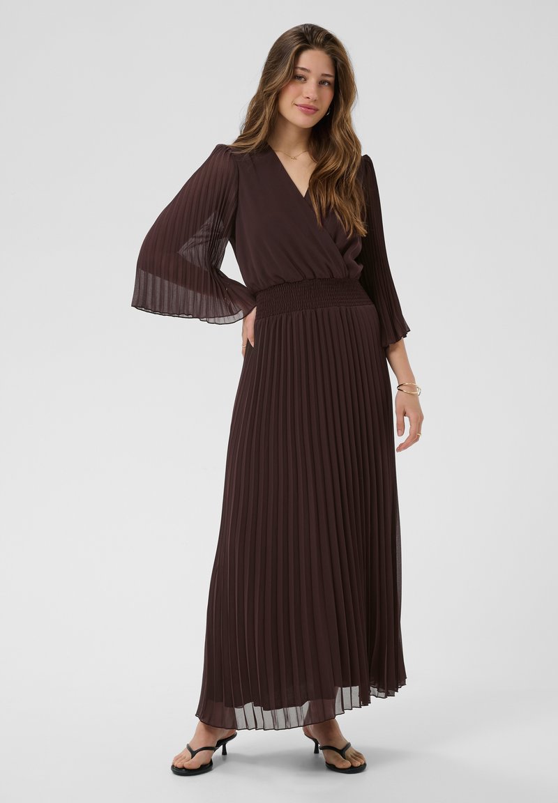 Femme portant une longue robe plissée marron foncé avec des manches cloche transparentes et des sandales à talons noires, debout devant un fond clair uni.