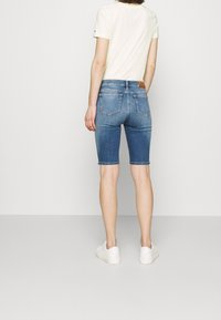 Džínové šortky v středně modré barvě, s těsným střihem a hladkým povrchem. Zadní kapsy s jemným prošíváním a hnědá kožená nášivka v pase.