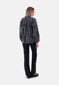 Donna che indossa una blusa nera a motivi paisley e pantaloni neri, in piedi con la schiena rivolta e la testa leggermente inclinata di lato.