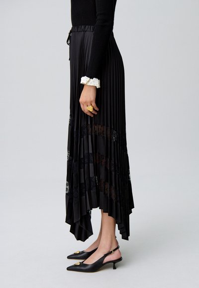 Jupe plissée noire avec ourlet asymétrique et panneaux en dentelle. Associée à des chaussures à bride arrière noires avec un accent doré.