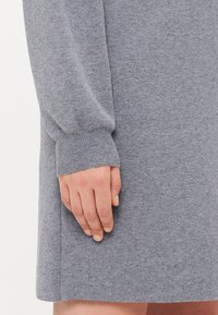 Grå strikket kjole med lange ermer, ribbestrikkede mansjetter og en myk tekstur. Nærbilde av en hånd som hviler på stoffet, som fremhever materialets veving.