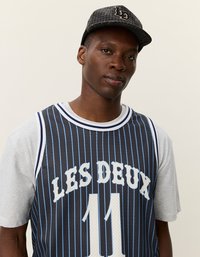 Fekete és kék csíkos kosárlabda mez "LES DEUX" felirattal és 11-es számmal, egy szürke rövidujjú póló fölöttrétegezve. Texturált sapka a fején.