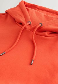 Svetlooranžová mikina s kapucňou s ladnými šnúrkami, ktoré majú kovové zakončenia a čierne oká na jednoltivom pozadí.