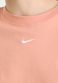 Peach cotton T-shirt featuring a ribbed crew neckline and a white embroidered Nike swoosh logo on the left chest. Smooth texture.