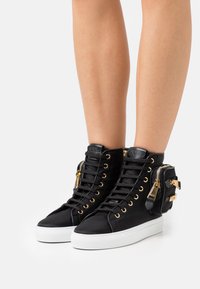 Svarta högklackade sneakers i tyg med gyllene öglor, en vit sula och en sidficka med dragkedja och hårdvarupynt.