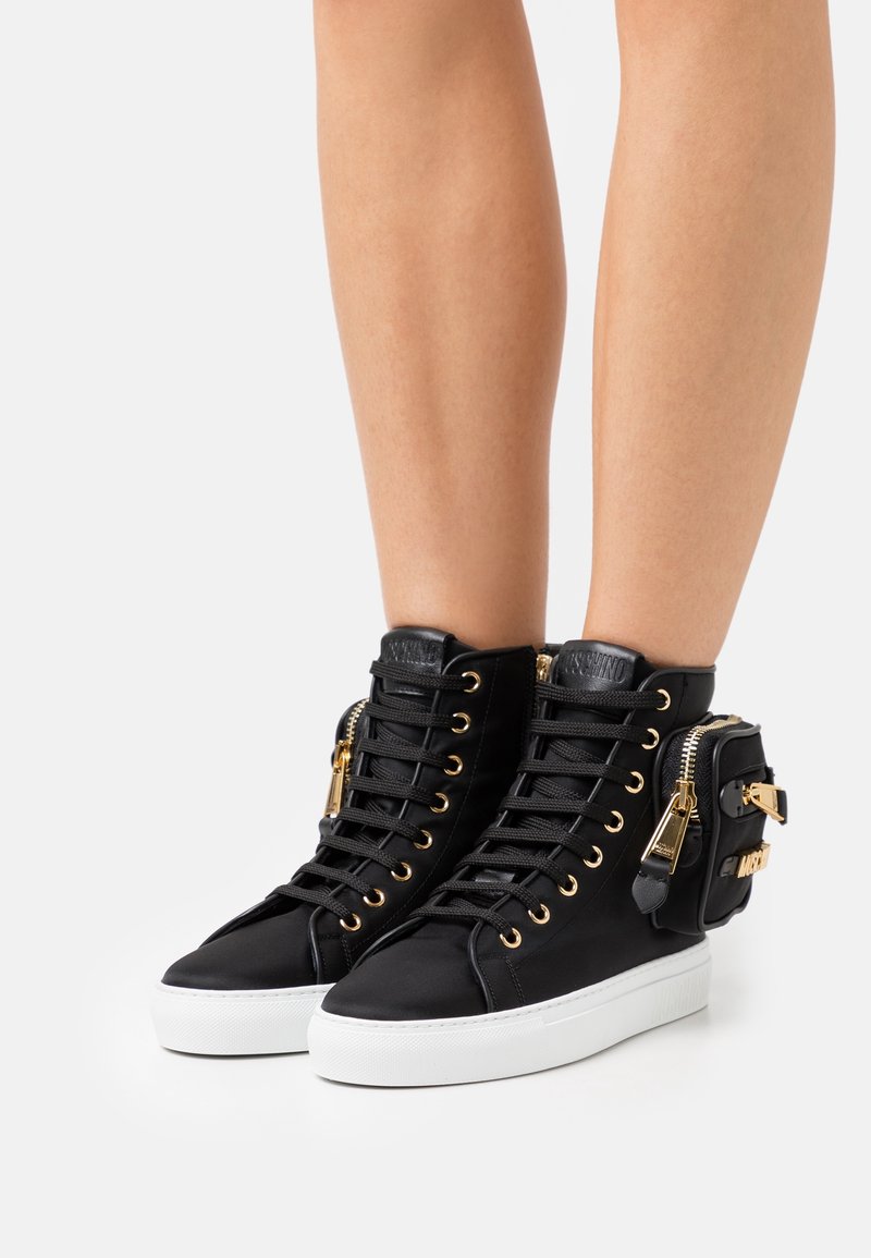 Svarta högklackade sneakers i tyg med gyllene öglor, en vit sula och en sidficka med dragkedja och hårdvarupynt.