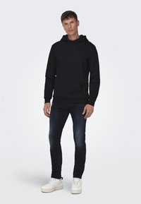 Hanorac negru cu buzunar tip cangur și șnururi, purtat cu blugi închiși la culoare și adidași albi. Design simplu și textură moale.