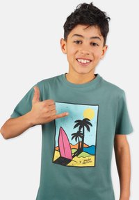 Vihreä puuvillapaita, jossa on värikäs surf-kohtauskuva, jossa on vaaleanpunainen purjelauta, palmuja ja aurinko rannikkotaustalla.