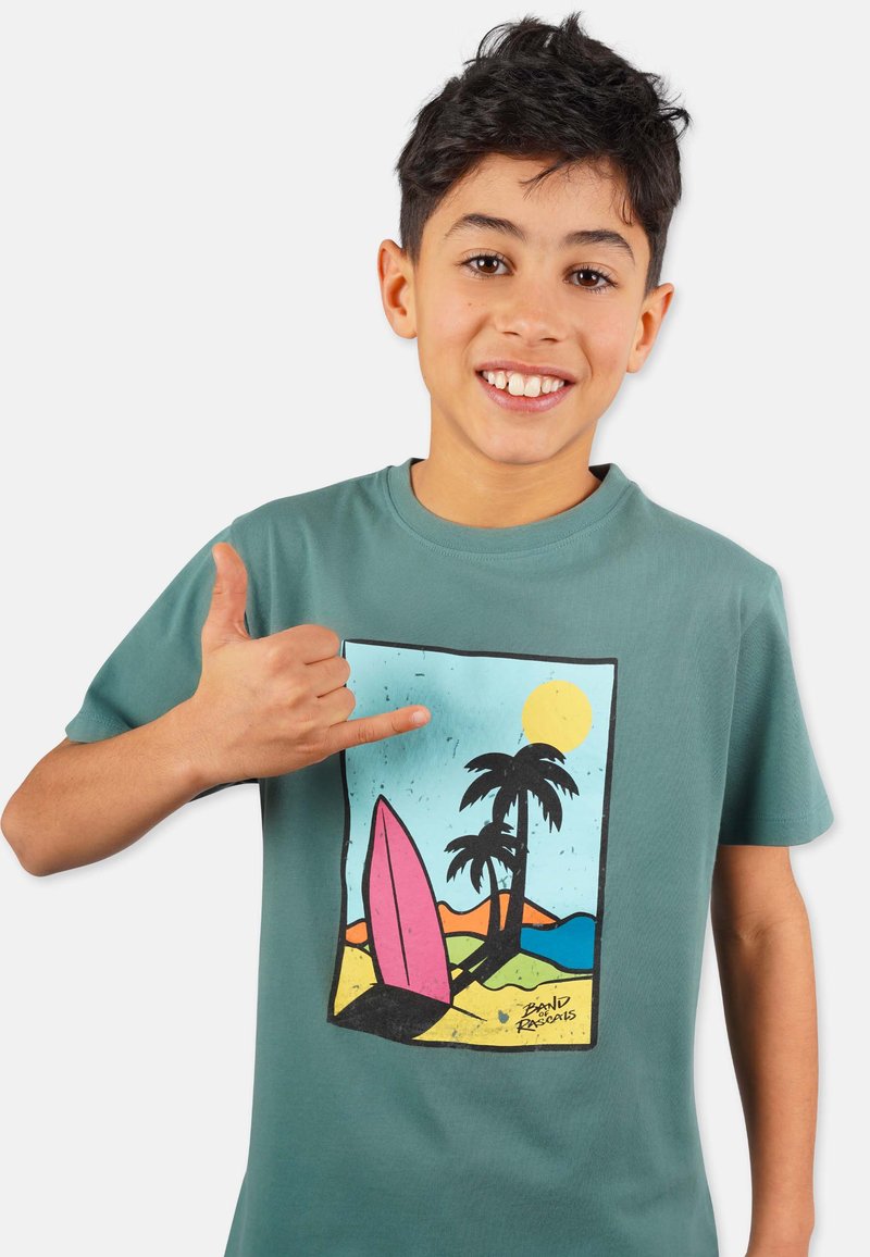 Vihreä puuvillapaita, jossa on värikäs surf-kohtauskuva, jossa on vaaleanpunainen purjelauta, palmuja ja aurinko rannikkotaustalla.