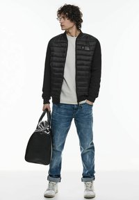 Svart puffjacka med ribbade ärmar, grå skjorta, ljusblå jeans och vita sneakers. Bär en svart väska med glänsande yta.