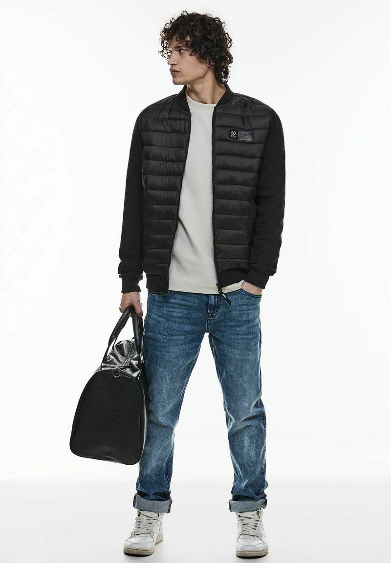 Svart puffjacka med ribbade ärmar, grå skjorta, ljusblå jeans och vita sneakers. Bär en svart väska med glänsande yta.