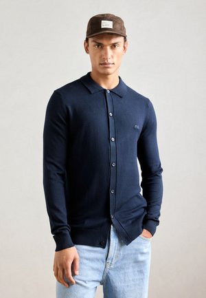 Navy blue long-sleeve cardigan, buttoned front, ribbed cuffs, smooth texture, worn with light blue jeans, featuring a logo on the left chest.