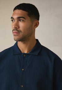 Tengerészkék, rövid ujjú ing strukturált anyagból, jellegzetes gallérral, három gombbal az elején és laza szabással.