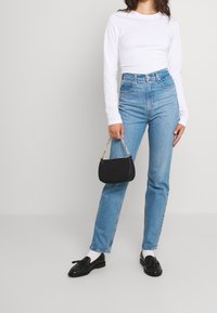 Blusa blanca de manga larga, jeans de tiro alto en un azul claro, bolso negro con cadena dorada, zapatos negros. El modelo está de pie en un fondo neutro.