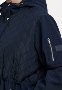 Navy quilted jacket with a hood, featuring a smooth texture, zippered pocket on the sleeve, and an embossed logo on the chest.