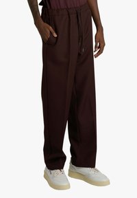 Pantaloni jogger color marrone scuro con vita elasticizzata, coulisse, due tasche laterali e vestibilità ampia, abbinati a sneakers bianche.