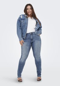 Chaqueta de mezclilla con acentos desgastados, combinada con una camiseta blanca y unos jeans ajustados de color azul claro. Sandalias negras completan el conjunto.