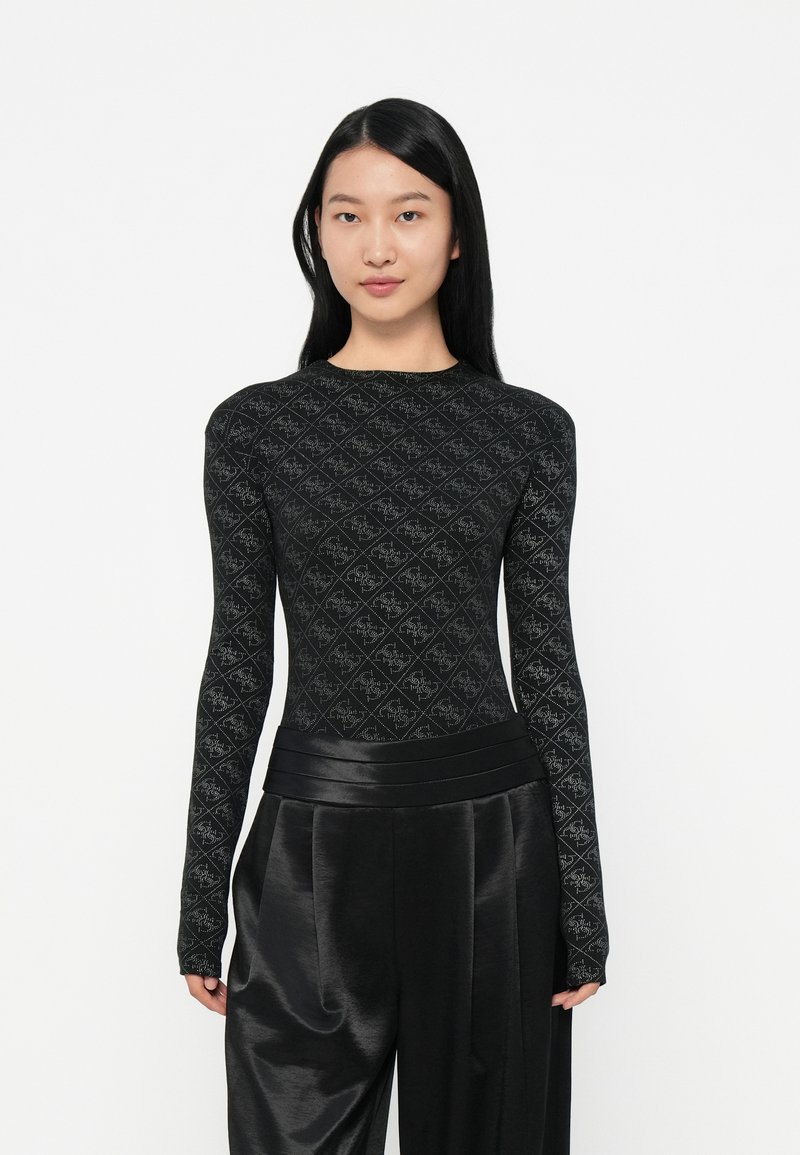 Una donna con lunghi capelli neri che indossa un top a maniche lunghe con motivo nero e pantaloni a vita alta lucidi neri su uno sfondo bianco semplice.