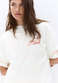Hvid oversized t-shirt med koralfarvet kursiv "Stvd" tekst, korte rullede ærmer med stribede accenter og en glat overflade. Sølv kæde halskæde.