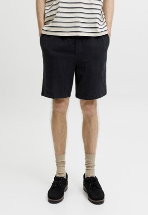 Person iført sorte teksturerede shorts, beige strømper og sorte snøresko står med hænderne i lommerne mod en ensfarvet baggrund.