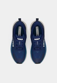 Lichte blauwe hardloopschoenen met een bovenwerk van mesh, voorzien van een ronde neus, een gevoerde kraag en een rubberen zool. Het 'HOKA'-logo is goed zichtbaar.