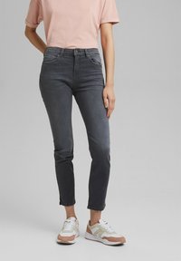 Persona vistiendo jeans ajustados de tiro alto en gris oscuro, una camiseta de manga corta en rosa claro y zapatillas blancas con detalles en rosa, de pie contra un fondo gris.