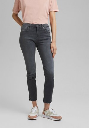 Person som bär höga, mörkgrå skinny jeans, ljusrosa kortärmad skjorta och vita sneakers med rosa detaljer, stående mot en grå bakgrund.