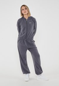 Tuta in velour grigio con felpa con zip e polsini elastici, con accenti di strisce laterali e il logo "EA7" sul petto. Sneakers bianche.