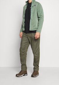 Chaqueta de forro polar verde de Patagonia con cuello alto, camiseta negra, pantalones texturizados en verde oliva y botas de senderismo marrones con suelas blancas.