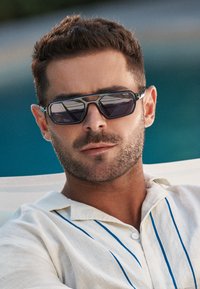 Homme aux cheveux courts et bruns avec une barbe taillée portant de grandes lunettes de soleil rectangulaires et une chemise blanche à rayures bleues.