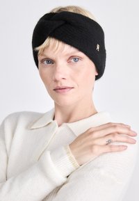 Fascia in maglia nera con design intrecciato, impreziosita da un emblema dorato; il modello indossa un maglione in maglia leggera e dalle texture chiare.