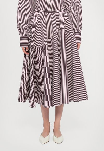Jupe rayée en blanc et rouge foncé, avec un design asymétrique à plusieurs couches. Fabriquée en tissu léger, arrivant au genou, associée à des chaussures crème.