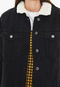 Veste en denim noir avec un col en shearling blanc, fermeture à boutons sur le devant et poches poitrine ; portée par-dessus une chemise à carreaux jaune et noire.